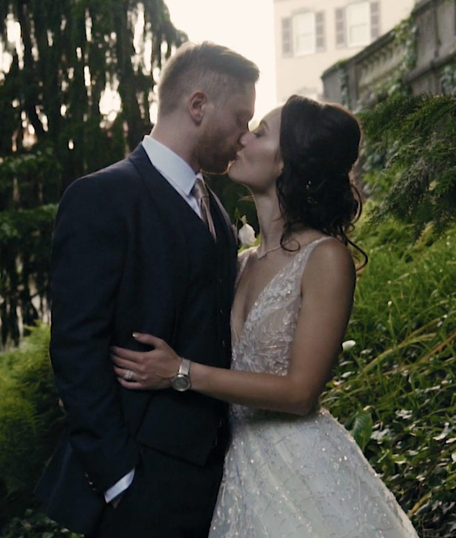A couple in formal attire shares a tender kiss outdoors, surrounded by greenery and trees at their picturesque Winterthur wedding.